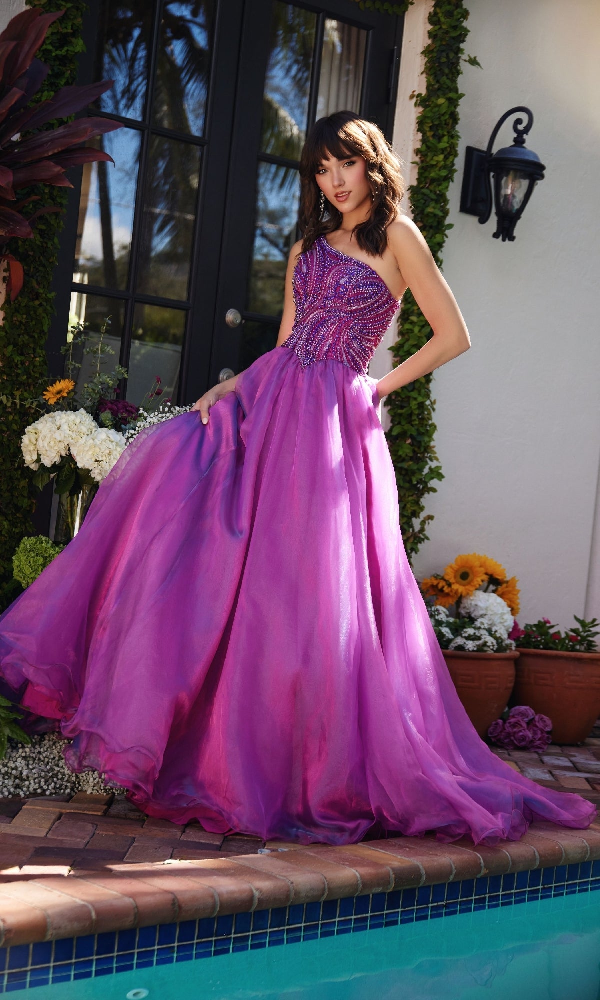 A woman in the Ava Presley 29258 one-shoulder prom ball gown stands by a poolside garden, surrounded by flowers and greenery, gently holding the side of her dress.